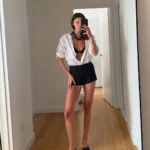 Mulher em frente ao espelho vestindo uma camisa branca feminina modelo oversized, usada aberta com top e minissaia preta, compondo um look despojado e elegante.