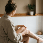 Mulher de costas vestindo um roupão neutro aplicando creme hidratante nas pernas em um banheiro com luz natural, ilustrando um passo essencial da rotina corporal de autocuidado.