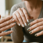 Close-up de três mulheres de diferentes gerações exibindo tendências de unhas 2026. Uma mão tem unhas amendoadas nude brilhante, outra unhas quadradas metálicas prateadas e a terceira unhas curtas verde oliva com acabamento fosco. A imagem destaca a diversidade de estilos e acabamentos que serão tendência em 2026.